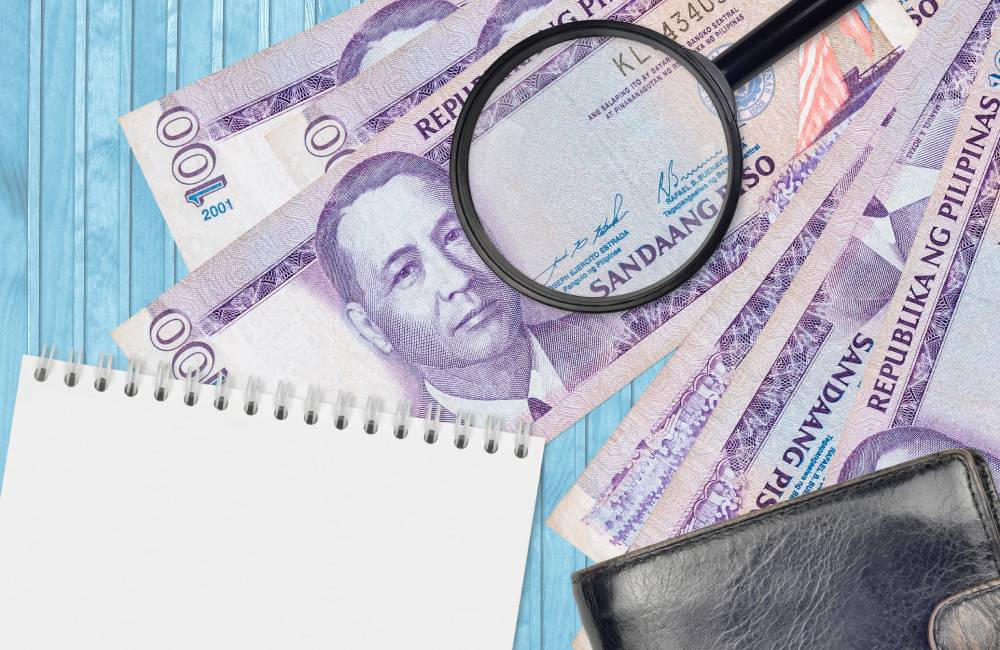 Magnifying glass lying on top of Philippine pesos showing that the BIR will assess late fees on an Extrajudicial Settlement that is submitted more than 1 year after the deceased's death.