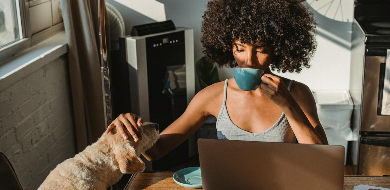 A woman sipping her morning coffee while petting her dog in front of her computer